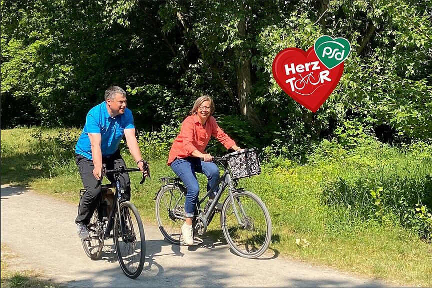 Eine Radfahrerin und ein Radfahrer radeln auf einem naturbelassenem Weg durch eine baumreiche Gegend. Ein rotes Herz mit der Inschrift HerzTour, wobei das ou durch ein Fahrrad symbolisiert wird.