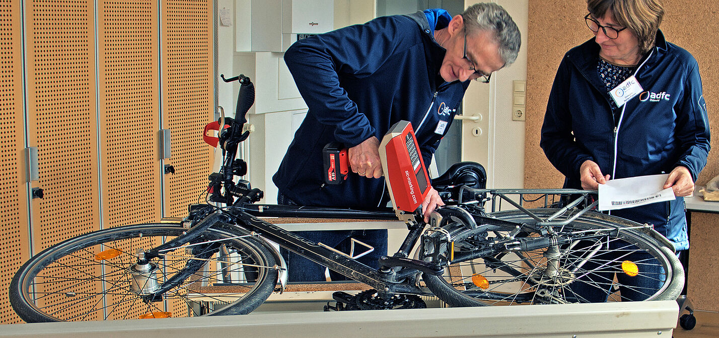 Fahrrad-Codierung Nadeldrucker Ein Mann codiert mit einem Nadeldrucker ein auf der Seite liegendes Fahrrad, während die Besitzerin zuschaut.