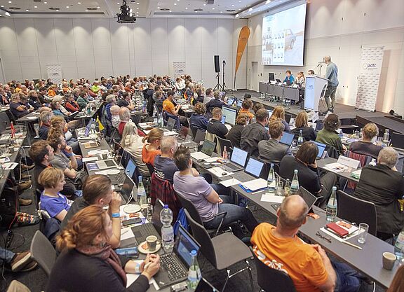 von Heesen Ein grpßer Saal mit 140 Menschen an Tischreihen, die zur Bühne blicken, wo Boris von Heesen einen Vortrag hält.
